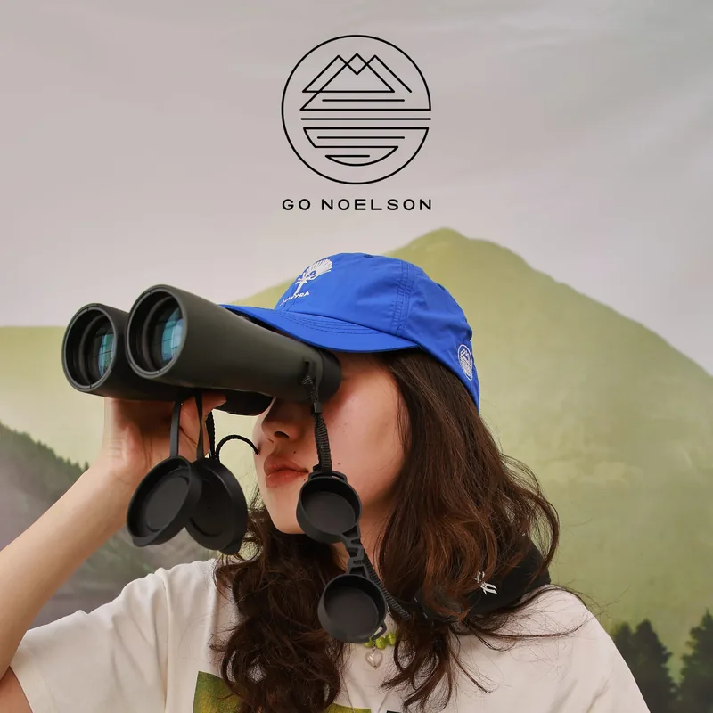Woman wearing a blue Go Noelson cap looks through binoculars in the mountains of Kyrgyzstan